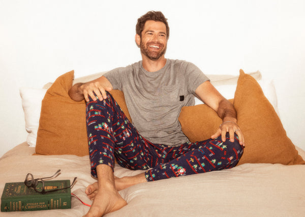 Man sitting in bed while wearing gray tee and navy blue Sleep bottoms in a holiday print